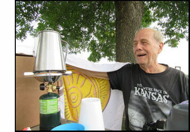 Picture shows David sitting to the right of a campstove with a large coffee pot on it.  He is smiling and talking, and reaching his arm out to a tall box to the left of the coffee pot.  A towel draped on his arm extends from his body to the box. 
